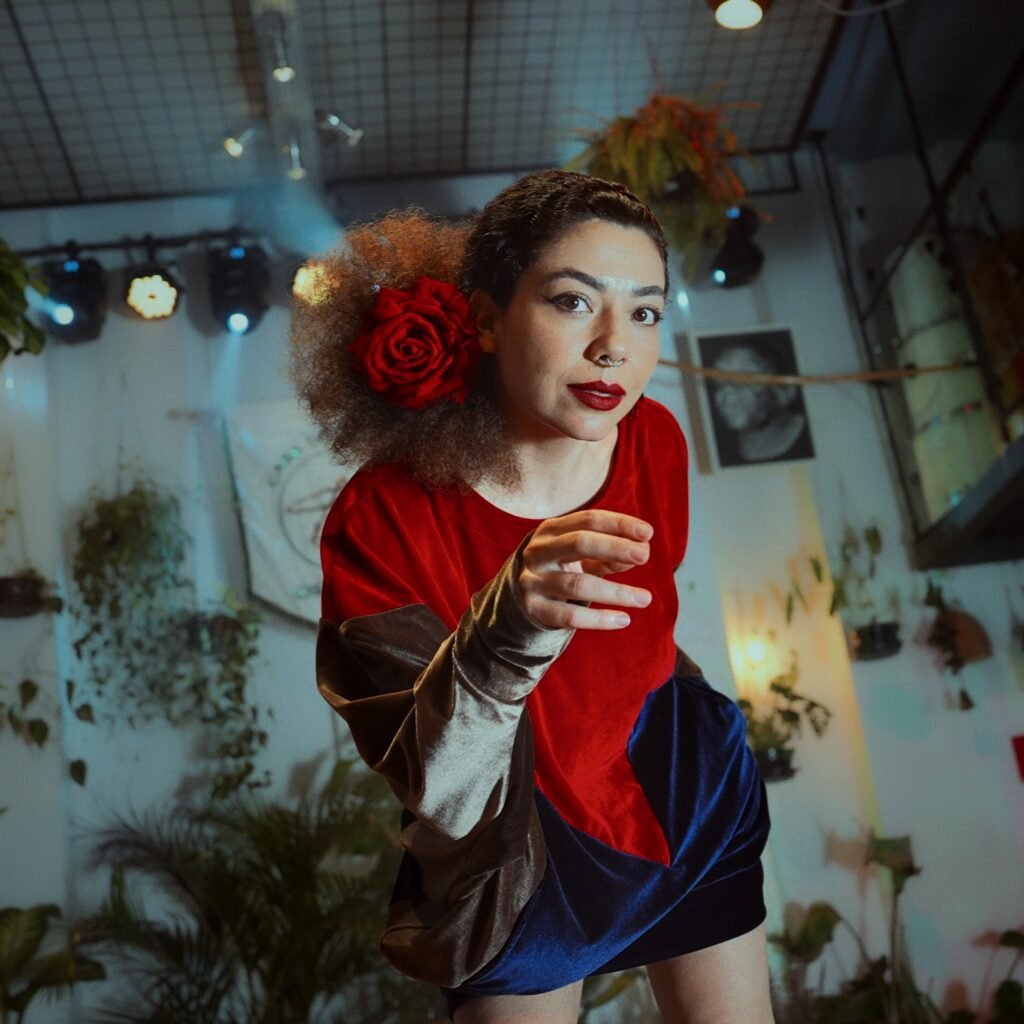 Fotografia promocional em enquadramento quadrado da cantora Vanessa Moreno em perspectiva de ângulo baixo. Ela se inclina em direção à câmera estendendo a mão direita, enquanto olha fixamente para a lente. Veste uma peça tricolor de veludo (vermelho, azul e bege) e traz uma grande rosa vermelha presa nos cabelos cacheados presos para trás. Ao fundo, veem-se refletores de palco, plantas suspensas e um quadro na parede sob iluminação suave.