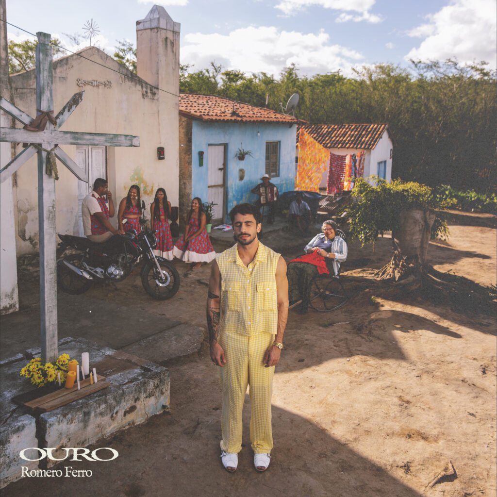 Capa do álbum 'Ouro' de Romero Ferro. O cantor aparece em primeiro plano vestindo um conjunto amarelo xadrez, de olhos fechados, em um cenário rural com casas coloridas. Ao fundo, moradores locais, um sanfoneiro e mulheres vestidas com figurinos juninos compõem a cena